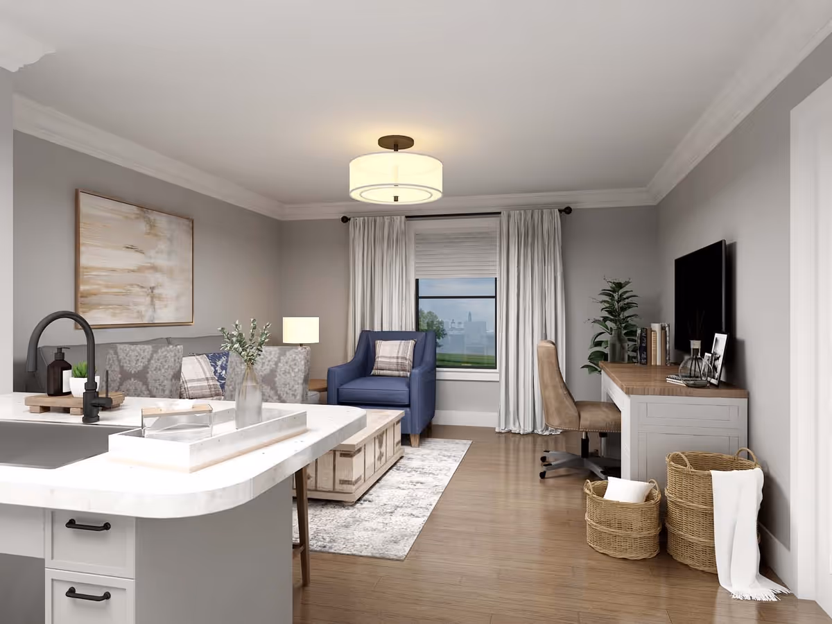 A modern living room and kitchen area with light gray walls and wooden flooring. The kitchen island has a white countertop with a black faucet and a small tray with decorative items. The living room features a blue armchair with a plaid pillow, a gray patterned sofa, a wooden coffee table on a light rug, and a window with white curtains. There is a desk with a brown chair, books, and decorative items next to a large flat-screen TV mounted on the wall. Two woven baskets with white pillows and a blanket are placed on the floor near the desk. A round ceiling light fixture illuminates the room.