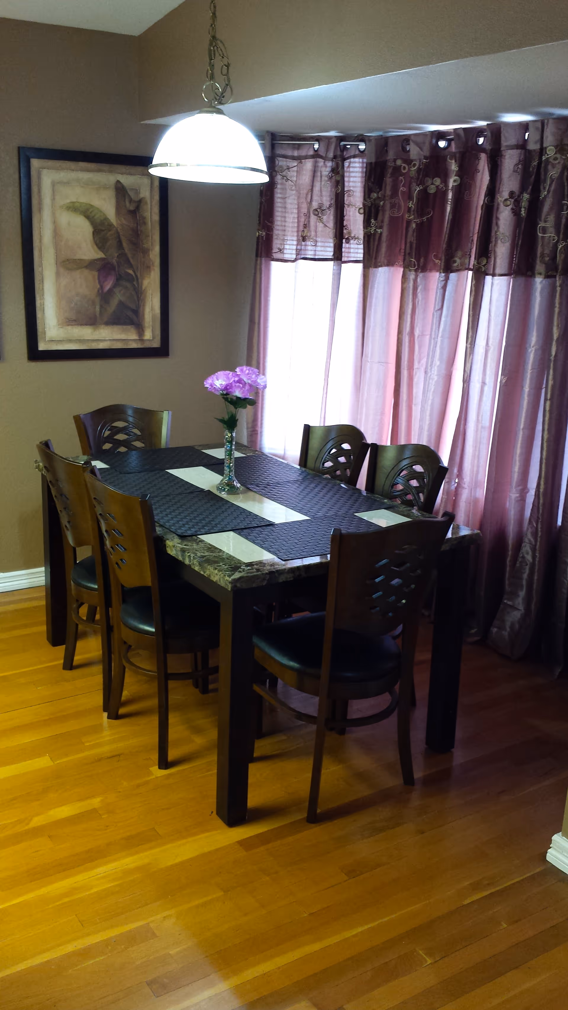 A dining room with a rectangular table covered with black placemats and a vase with purple flowers in the center. Six wooden chairs with black cushions surround the table. The room has wooden flooring, a hanging light fixture above the table, purple curtains covering the windows, and a framed botanical artwork on the wall.