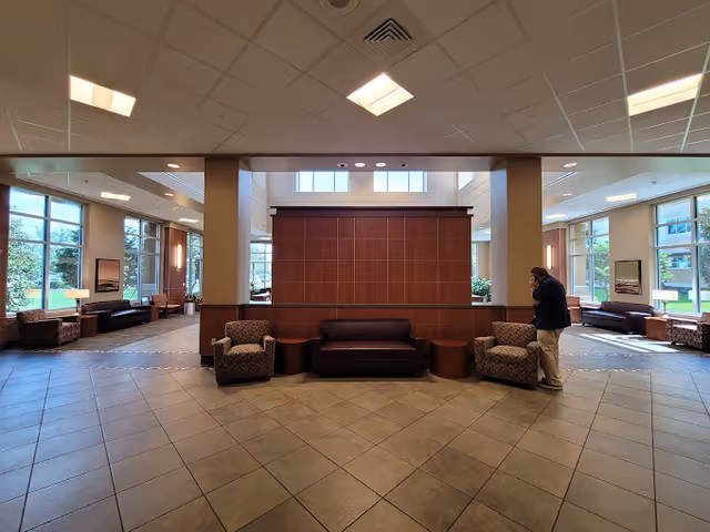 Spacious senior living facility common area with tiled floor, high ceiling, and large windows allowing natural light. The room features multiple seating arrangements including armchairs and sofas around wooden tables. A man is standing on the right side, talking on a phone. The walls have framed artwork and there are lamps providing additional lighting.