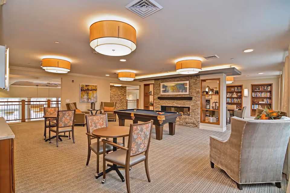 A spacious and well-lit common area in a retirement community featuring a pool table in the center, several round tables with floral upholstered chairs, a stone fireplace with a framed picture above it, built-in shelves with decorative items, and a cozy seating area with armchairs. The room has large ceiling lights and carpeted flooring.