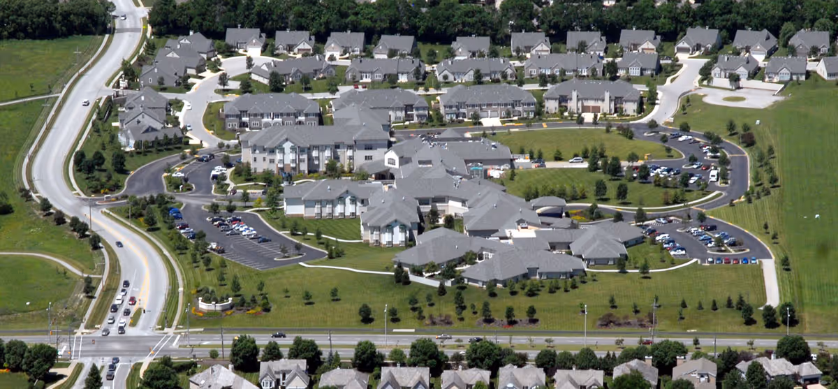 Aerial view of Village Shalom Senior Living in Overland Park showing multiple buildings with gray roofs, parking lots with cars, green lawns, and surrounding roads.