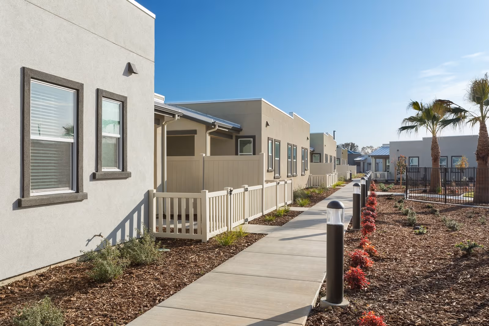 Paved walkway between landscaped beds leading past single-story beige senior living units with small fenced patios and palm trees under a clear blue sky.