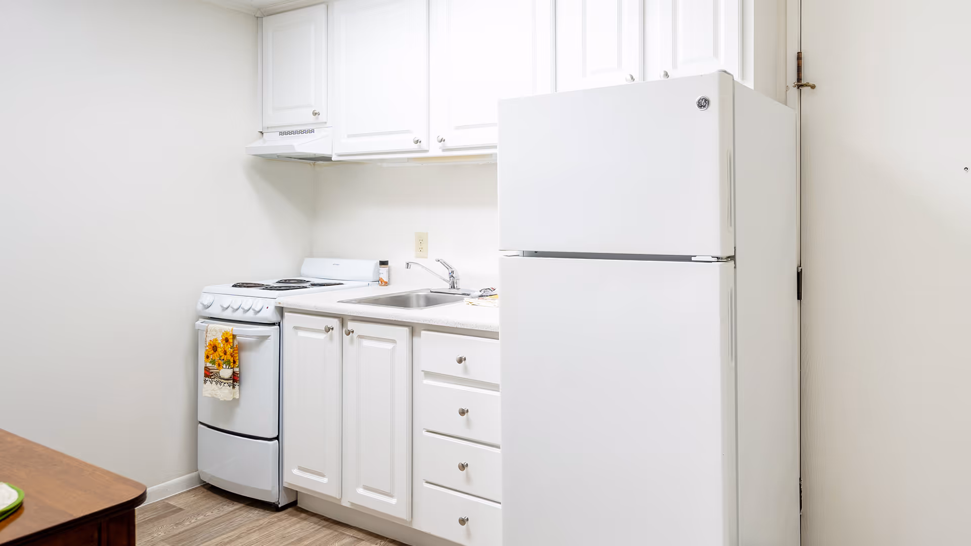 A small kitchen area with white cabinets, a white refrigerator, a white stove with a sunflower-themed towel hanging on the handle, a sink, and a wooden table partially visible on the left side.