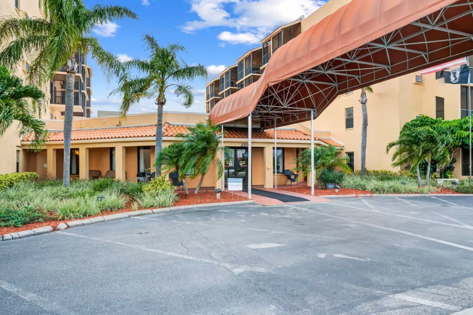 Front entrance of a stucco senior living building with a large maroon canopy, palm trees, landscaping, and a parking area.