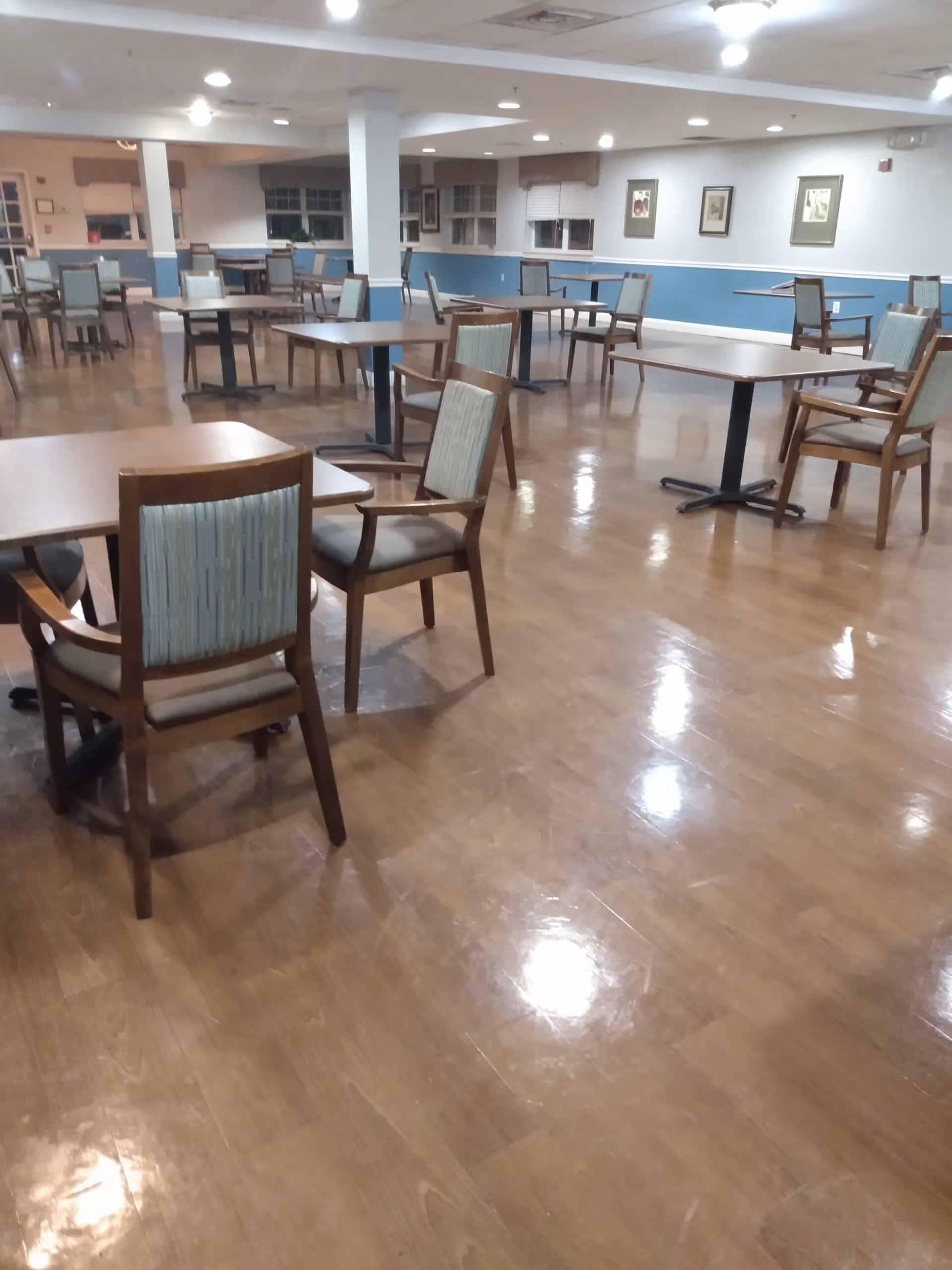 A spacious dining room with multiple wooden tables and chairs arranged neatly on a polished wooden floor. The walls are painted white with a blue lower half, decorated with framed pictures. The room is well-lit with ceiling lights and has windows with blinds.