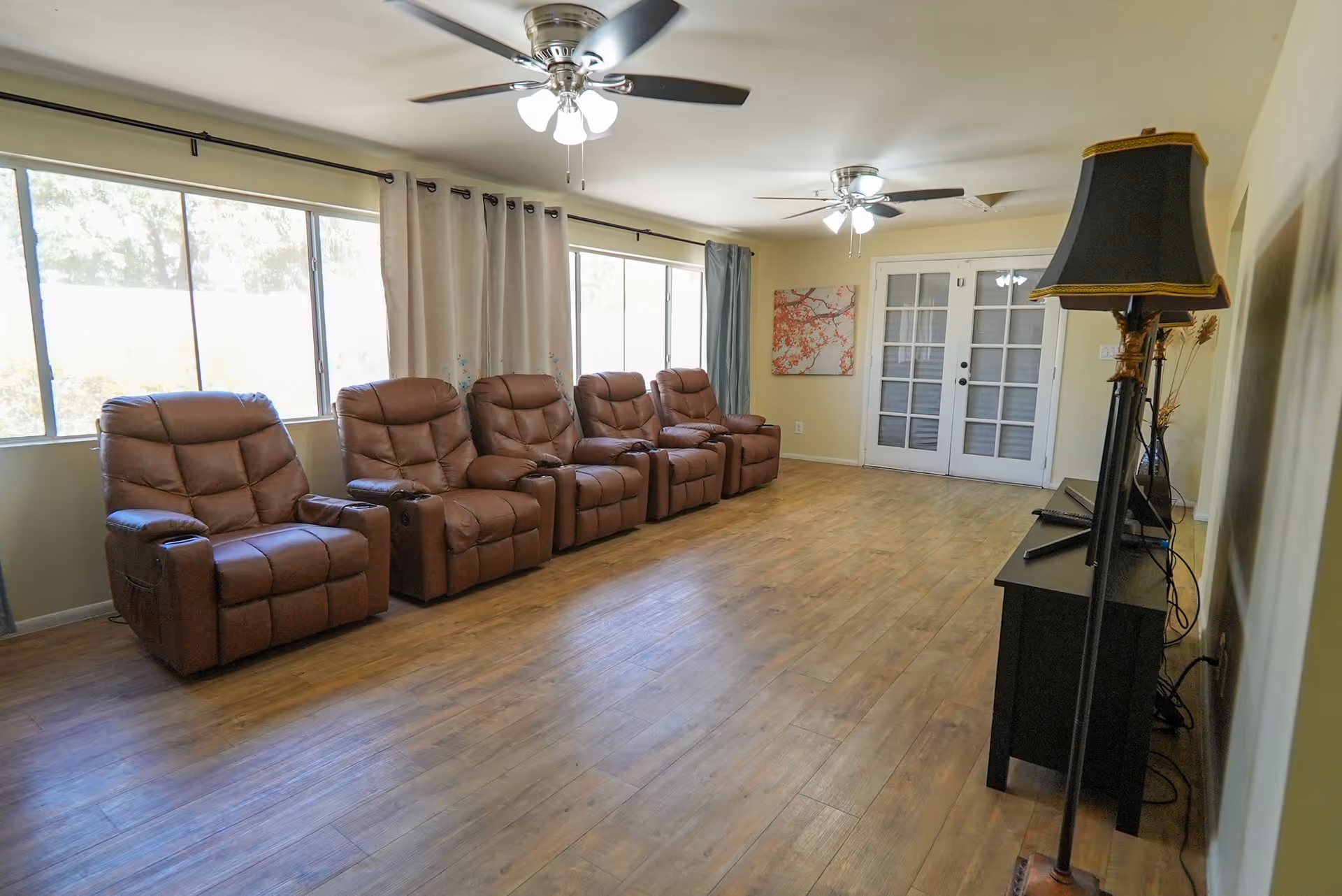 A living room with five brown leather recliner chairs lined up against a wall with large windows covered by curtains. The room has wooden flooring, two ceiling fans with lights, a black TV stand with a flat-screen TV, and a tall floor lamp with a black and gold lampshade. There is a painting on the far wall and double glass doors.