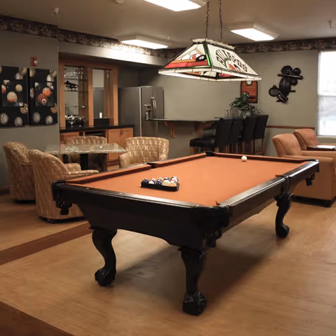 A senior living community recreation room with a pool table, seating area, and a small bar/kitchenette.