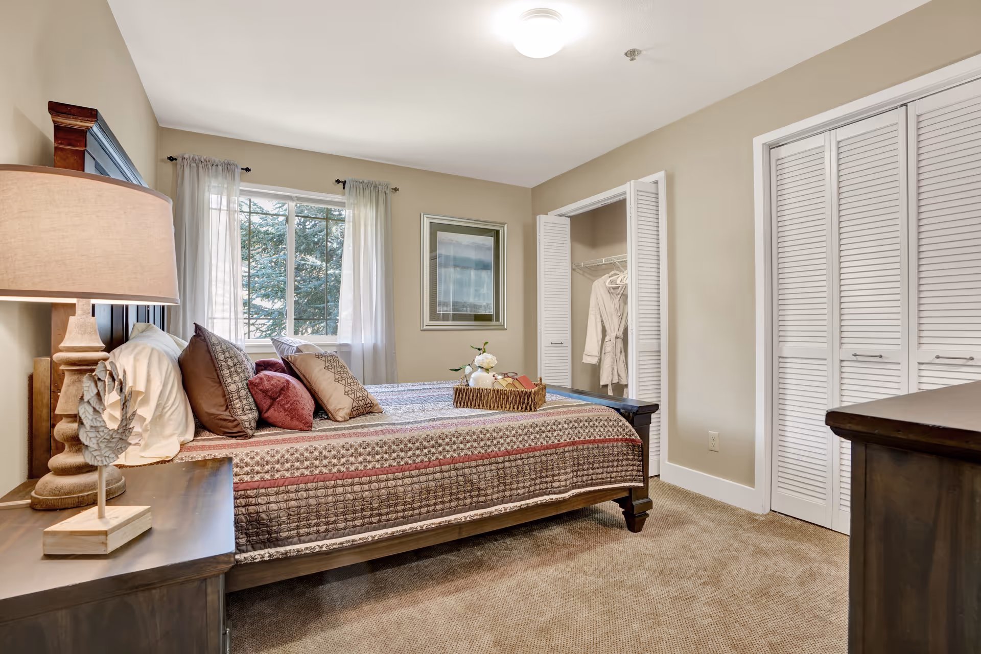 A cozy bedroom with a large bed covered in a patterned quilt and multiple pillows. There is a wooden nightstand with a lamp and decorative item on the left side of the bed. A window with sheer curtains lets in natural light. On the right side, there is an open closet with white louvered doors and a robe hanging inside. The walls are painted beige and the floor is carpeted.