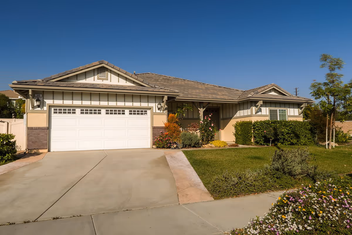 Single-story residential building with a two-car garage, beige and white exterior, a tiled roof, and a well-maintained front yard with green grass, bushes, and flowering plants under a clear blue sky.