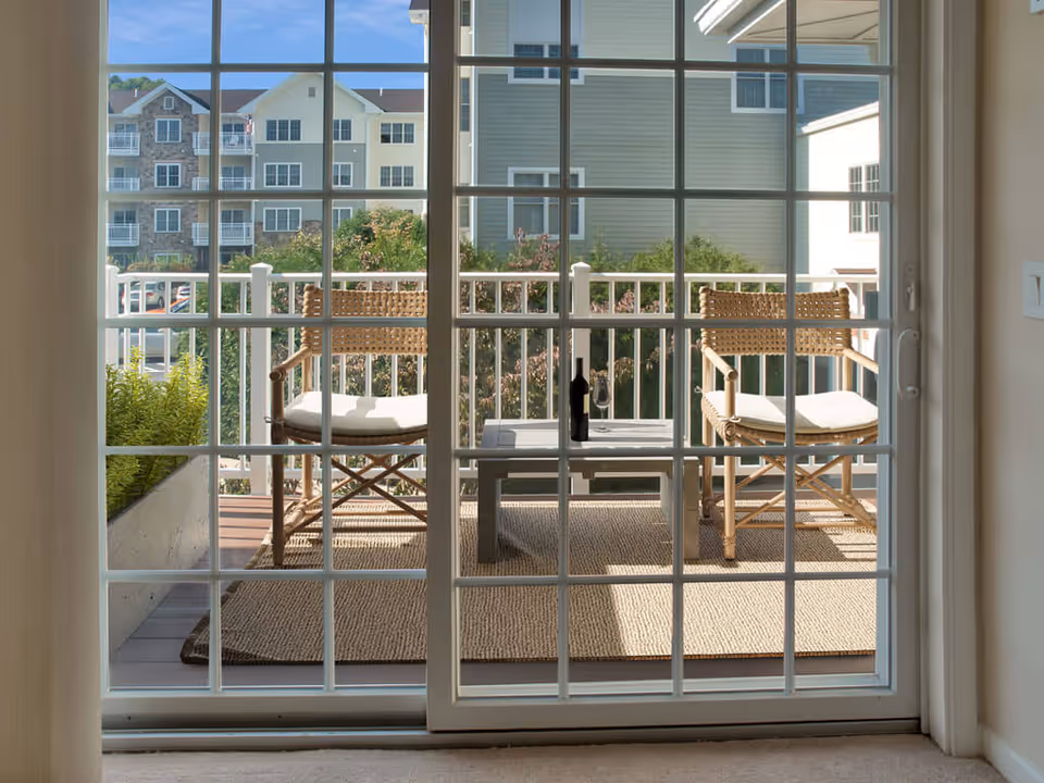 View through a large sliding glass door onto a small balcony with two wicker chairs with cushions and a small table with a bottle and glass on it. The balcony has a woven rug and a planter with greenery. In the background, there are multi-story residential buildings and trees under a clear blue sky.