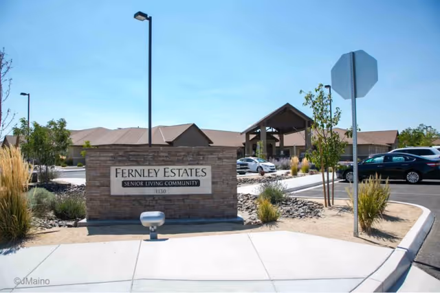 Entrance sign for Fernley Estates Senior Living Community with a stone base and surrounding landscaping, parking lot, and building with a covered entrance in the background under a clear blue sky.