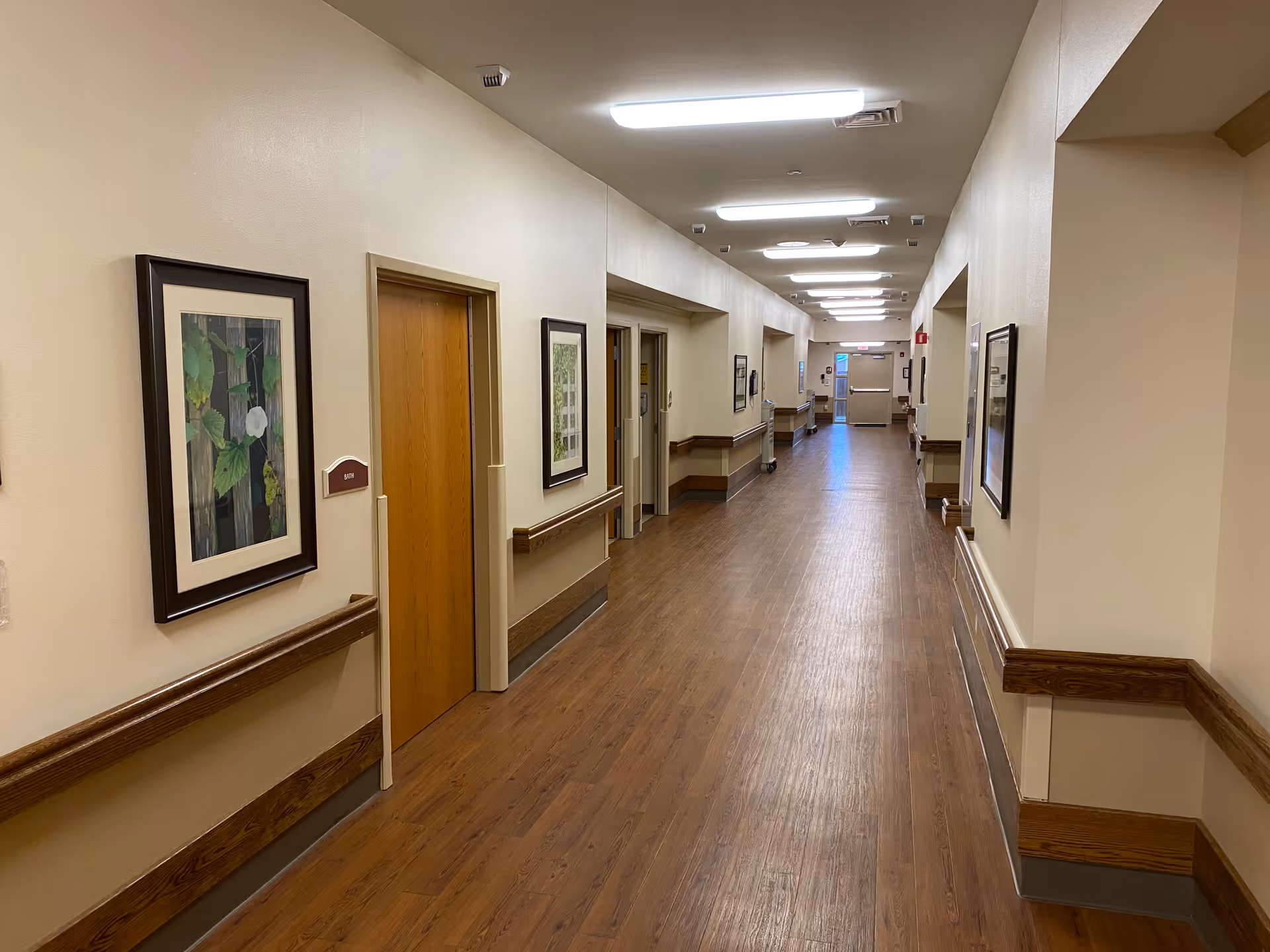 Well-lit long interior hallway with wood floors, handrails, doors, and framed artwork on the walls.