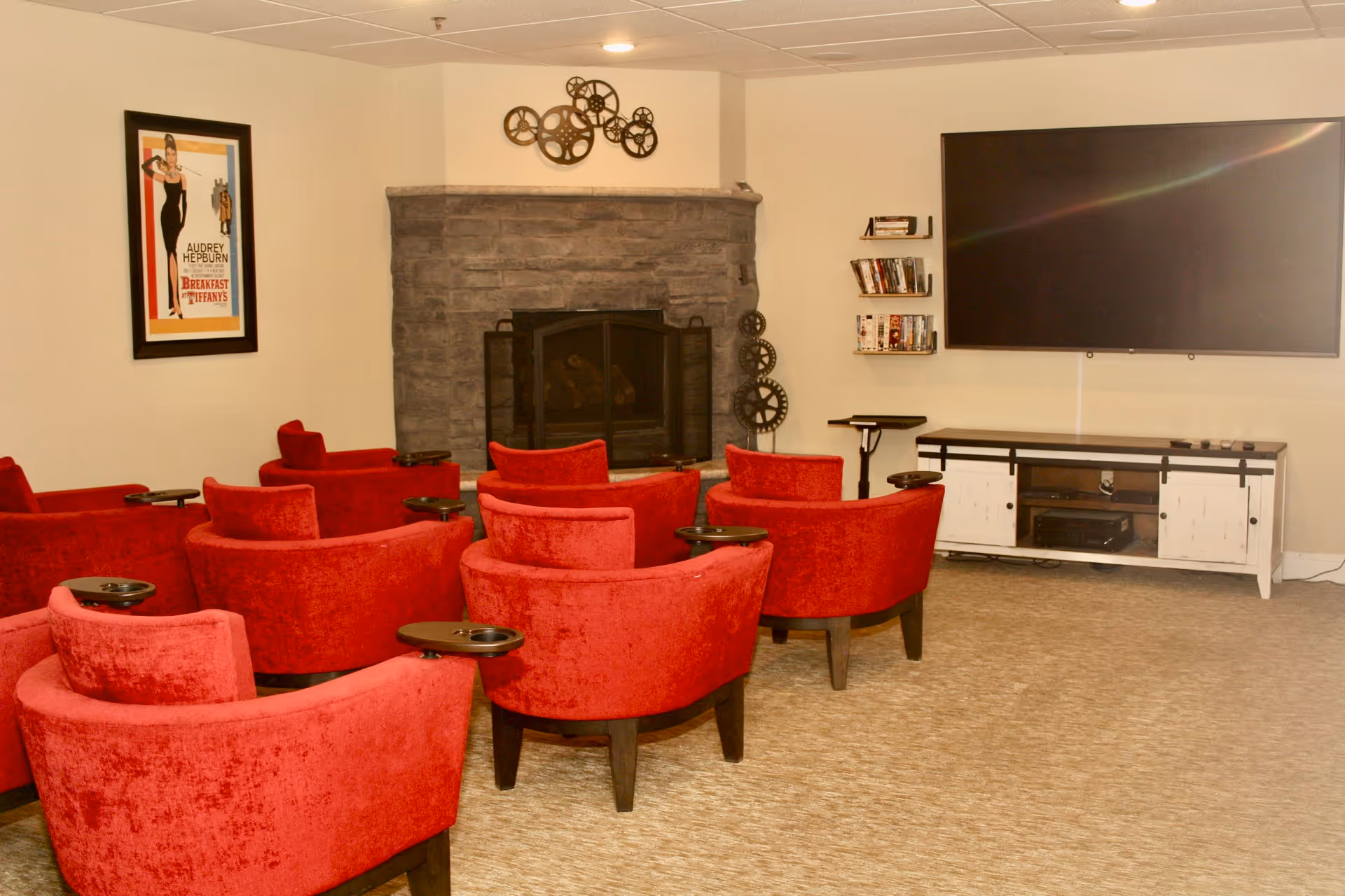 A cozy media room with several red upholstered armchairs arranged in rows facing a large flat-screen TV mounted on the wall. There is a stone fireplace with a decorative metal gear wall art above it. A framed poster of Audrey Hepburn from Breakfast at Tiffany's hangs on the left wall. Shelves with DVDs are mounted on the wall near the TV.
