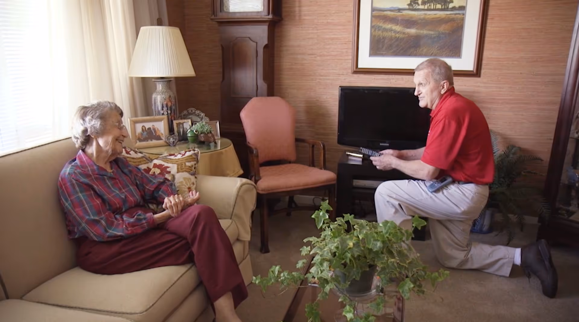 An elderly woman sitting on a beige couch smiling and talking to an elderly man kneeling on the floor holding a TV remote in a cozy living room with a lamp, framed photos, a television, and a potted plant on a glass table.