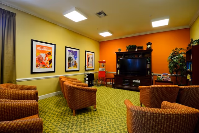 A cozy living room area with patterned carpet and orange accent wall. The room features six cushioned armchairs arranged in a semi-circle facing a large dark wood entertainment center with a flat-screen TV. On the entertainment center are decorative items including a clock and plants. The walls have framed artwork and there is a popcorn machine and a small table with board games on the right side. The room is well-lit with ceiling lights.