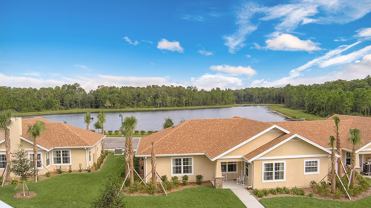 View of a senior living facility with beige buildings featuring brown shingle roofs, surrounded by green lawns and palm trees, with a large pond and forested area in the background under a blue sky with scattered clouds.