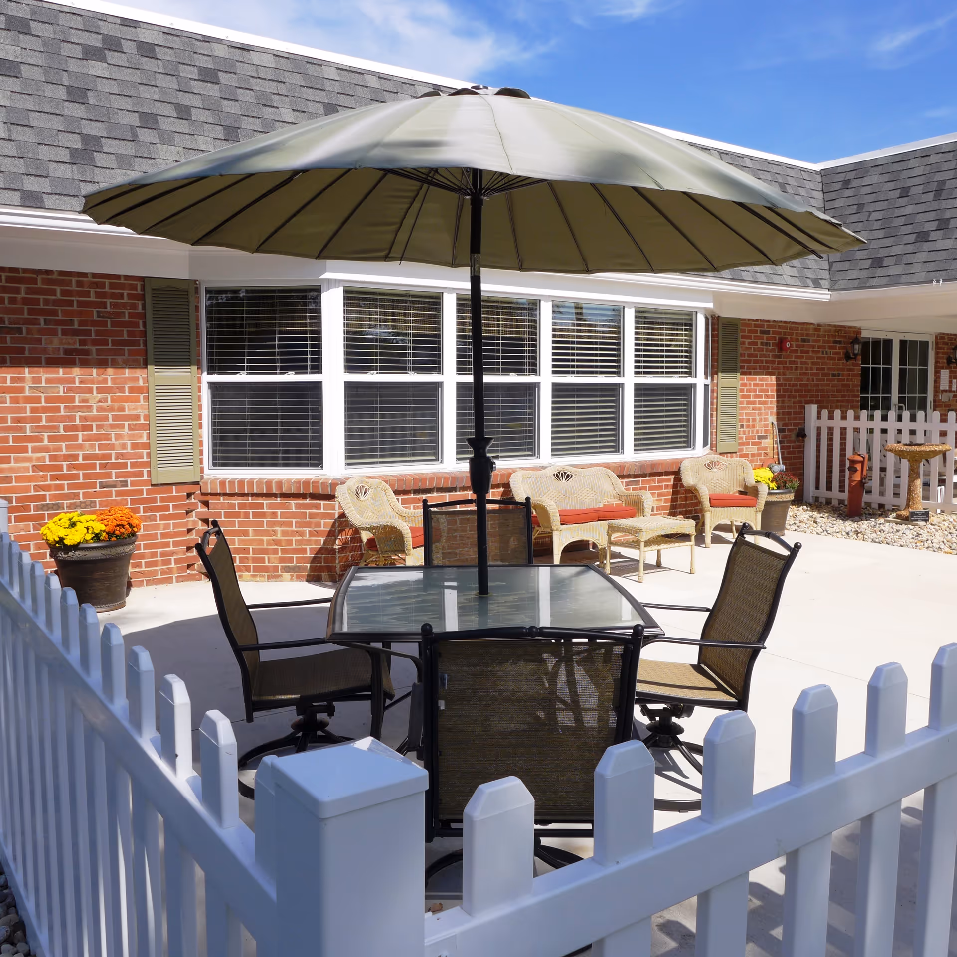 Outdoor patio area with a glass table and four chairs under a large green umbrella, surrounded by a white picket fence. In the background, there is a brick building with large windows and wicker seating with red cushions. Potted yellow and orange flowers are placed near the building.