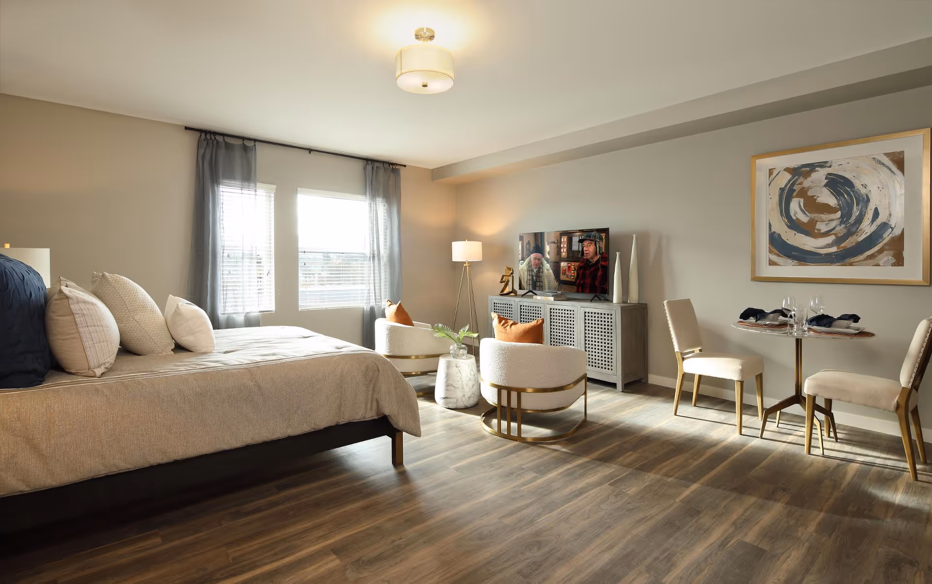 A spacious, well-lit room with a large bed on the left side, two modern armchairs with orange cushions around a small round marble table in the center, a TV on a cabinet against the far wall, and a small dining table with two chairs on the right. The room has wooden flooring, two windows with sheer curtains, a floor lamp, and abstract artwork on the wall.