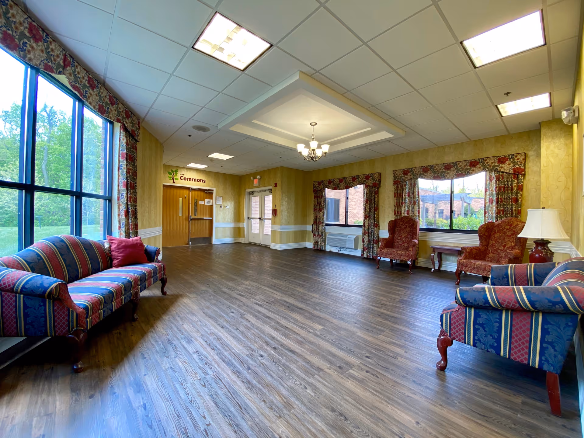 A spacious common area in a senior living facility with large windows letting in natural light. The room features patterned sofas and armchairs with floral curtains, wooden flooring, and a chandelier hanging from the ceiling. A sign above double doors reads 'Commons'.