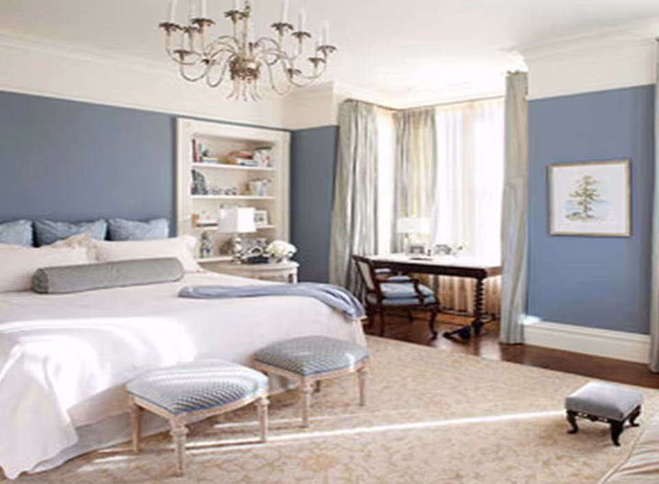 A bright and elegant bedroom with a large bed adorned with multiple pillows and a bolster cushion. Two upholstered stools are placed at the foot of the bed. The walls are painted blue with white trim, and there is a chandelier hanging from the ceiling. A built-in bookshelf with decorative items and a lamp is next to the bed. A wooden desk with a chair is positioned near large windows with light curtains, allowing natural light to fill the room. A framed picture hangs on the wall to the right.
