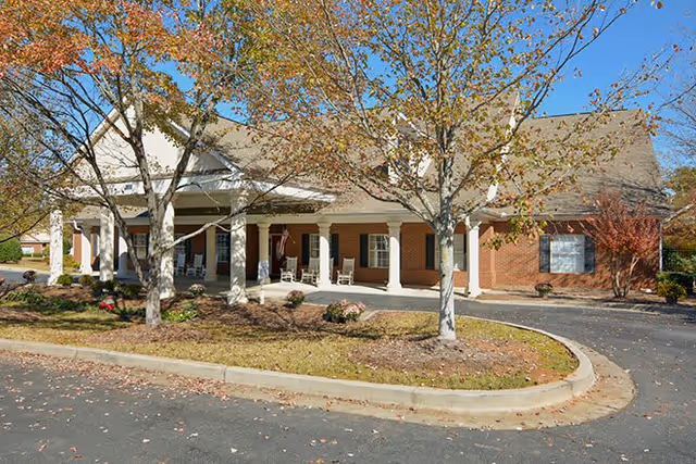 Exterior view of a single-story brick building with a covered porch supported by white columns. There are rocking chairs on the porch and trees with autumn foliage in front of the building. The sky is clear and blue.