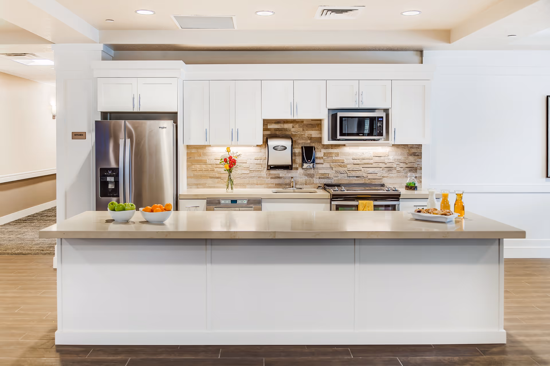 A modern kitchen with white cabinetry and a large island countertop. The kitchen features a stainless steel refrigerator, microwave, dishwasher, and stove. There are bowls of green apples and oranges on the island, along with a tray holding glasses of orange juice and a plate of cookies. A vase with flowers is placed on the counter against a stone tile backsplash.