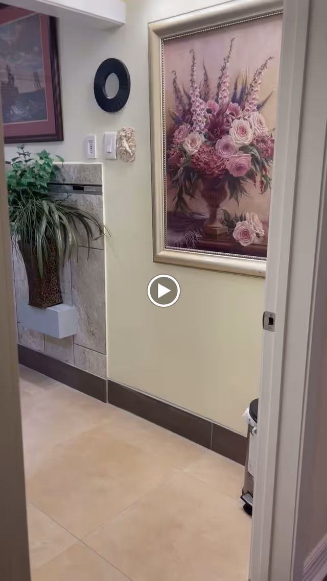 Interior hallway with beige tiled floor and light yellow walls. A large framed painting of a floral arrangement with pink and purple flowers hangs on the wall. There is a decorative black circular wall piece and a small sculpted face on the wall. A potted plant is placed on a tiled ledge to the left. A trash can is partially visible on the right side near the doorway.