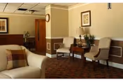 Comfortable sitting area in a senior living facility with chairs, a sofa, a side table and lamp, and framed artwork on the walls.