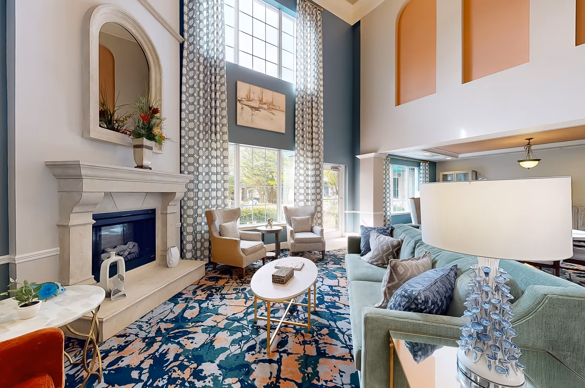 A bright and spacious living room with high ceilings and large windows covered by patterned curtains. The room features a light green sofa with decorative pillows, two beige armchairs around a small round table, a white oval coffee table, and a fireplace with a large mirror and a vase of flowers on the mantel. The walls are painted in soft blue and peach tones, and the floor is covered with a colorful patterned rug.