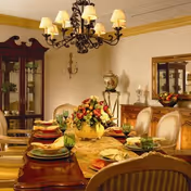 Elegant dining room with a large wooden table set for a meal, featuring plates, glasses, and a centerpiece of flowers. The room has upholstered chairs, a chandelier with lampshades, a glass-front cabinet, a mirror, and decorative items on a sideboard.