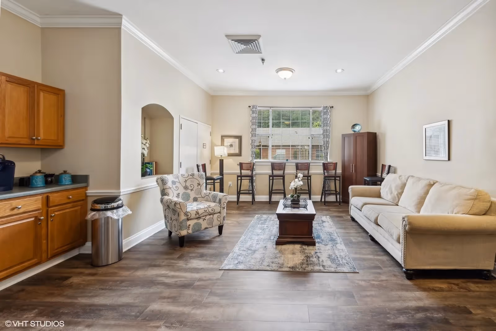 A bright and spacious living room area with a beige sofa on the right, a patterned armchair on the left, and a wooden coffee table on a rug in the center. Behind the coffee table, there is a window with four high chairs lined up against a counter. To the left, there are wooden kitchen cabinets and a trash can. The walls are painted light beige, and the floor is wood.