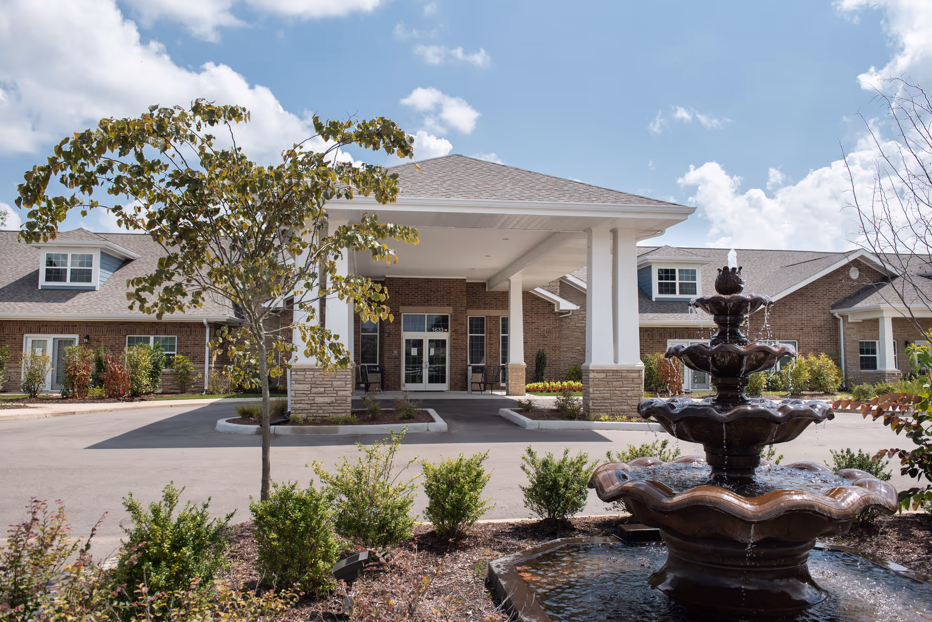 Front exterior view of a senior living facility with a covered entrance, brick walls, and a multi-tiered water fountain in the foreground surrounded by landscaping and small bushes under a partly cloudy sky.