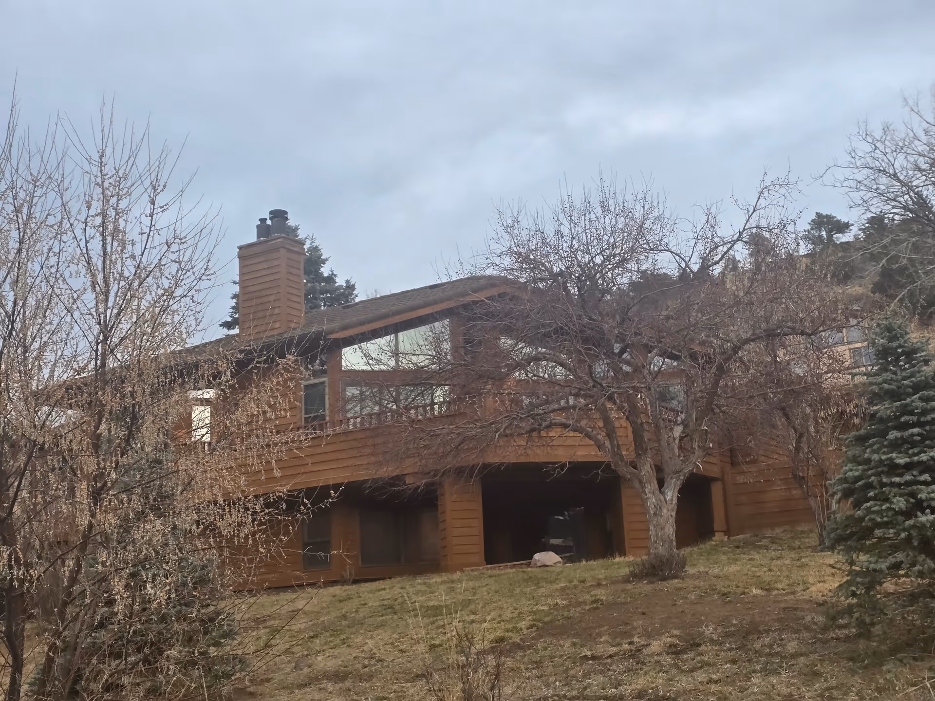 Two-story wooden building with large windows and a chimney set on a grassy slope with leafless trees in the foreground.