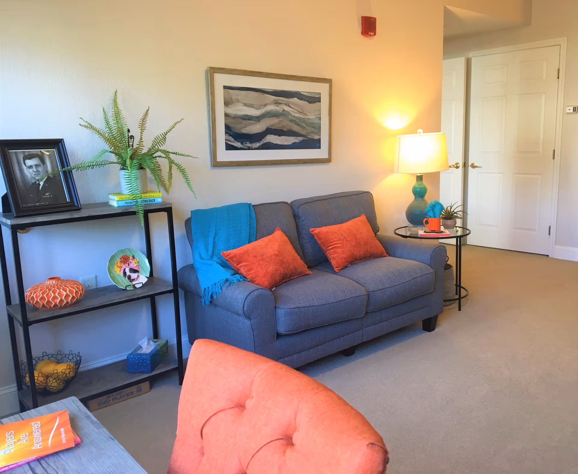 Bright living room with a blue loveseat topped with orange pillows and a turquoise throw, a side table with a lamp, shelving with decor, and wall art.