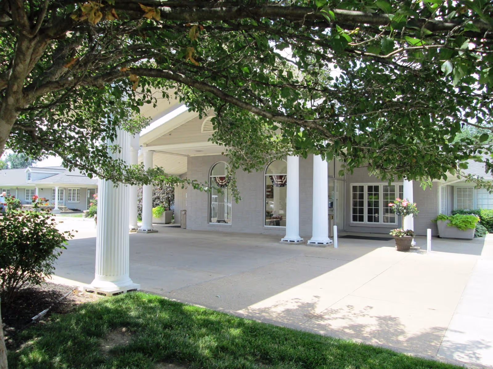 Entrance of a senior living facility with white columns supporting a covered driveway. The building has large windows and is surrounded by greenery, including a tree with dense foliage in the foreground and potted plants near the entrance.