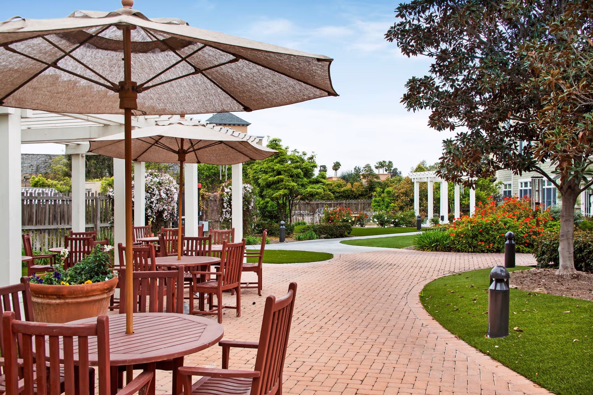 Outdoor patio area with round wooden tables and chairs under large beige umbrellas. The patio is paved with bricks and surrounded by green grass, trees, and colorful flowering bushes. A white pergola structure is visible in the background along with a wooden fence and a clear blue sky.