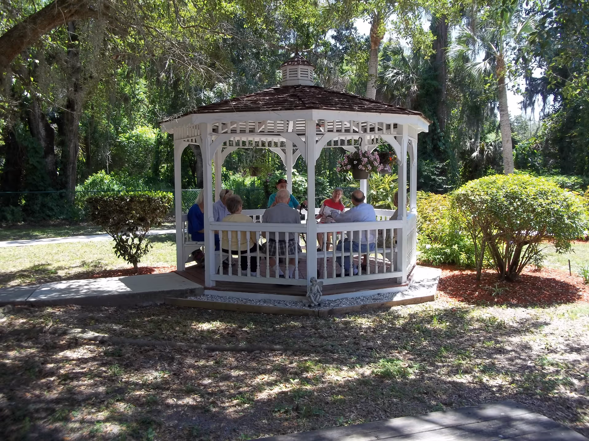 A group of elderly people sitting and conversing inside a white wooden gazebo in a garden area surrounded by trees and bushes on a sunny day.
