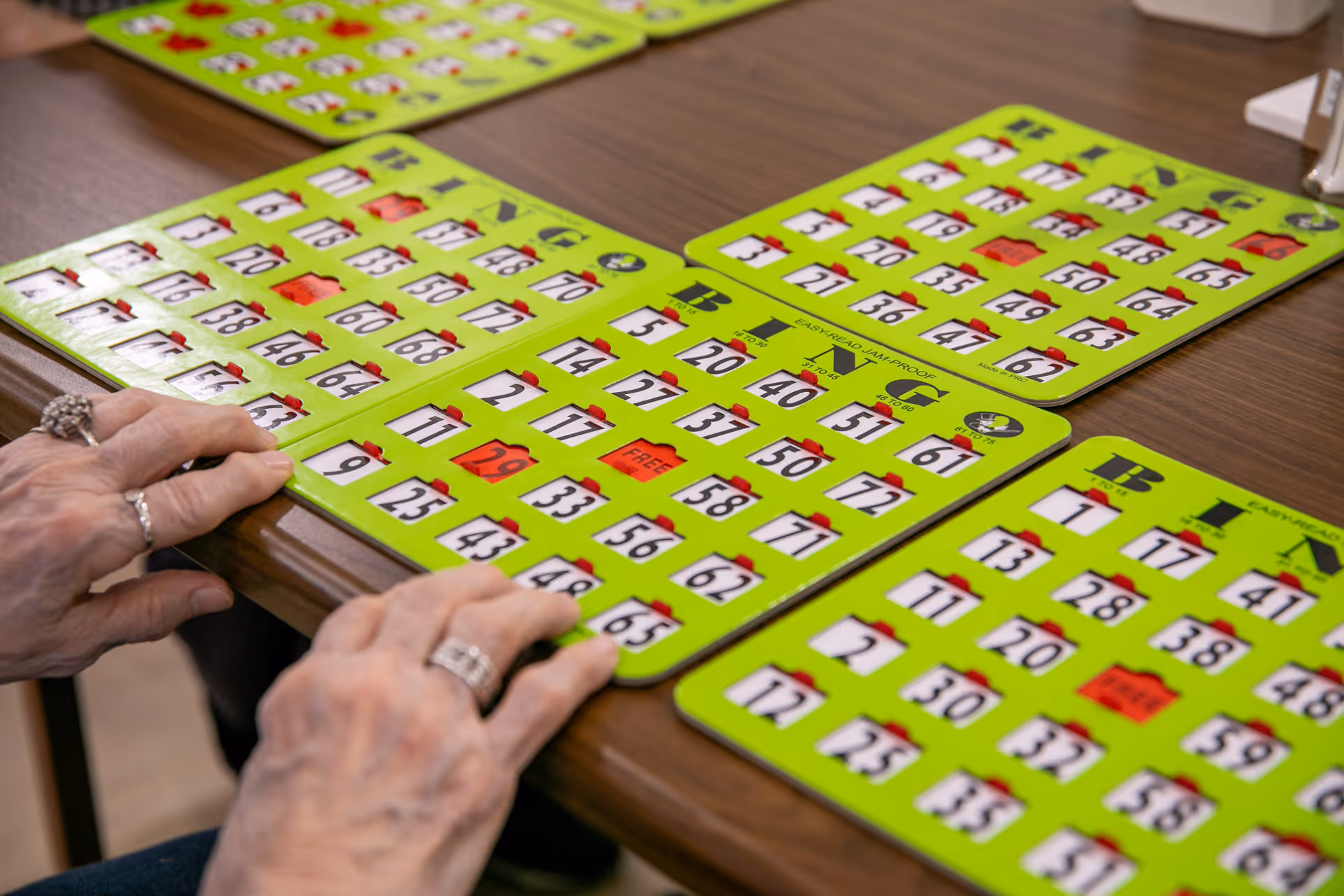 Close-up of an elderly person's hands with rings holding bright green bingo cards on a wooden table, with several other bingo cards visible in the background.