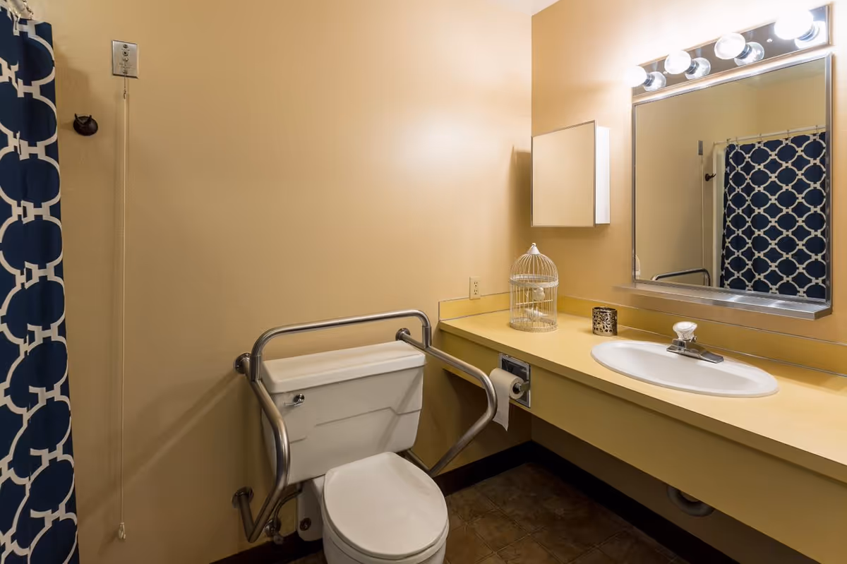 A bathroom with beige walls featuring a toilet with metal safety rails, a yellow countertop with a white sink, a large mirror with lights above it, a small decorative birdcage, and a blue and white patterned shower curtain.