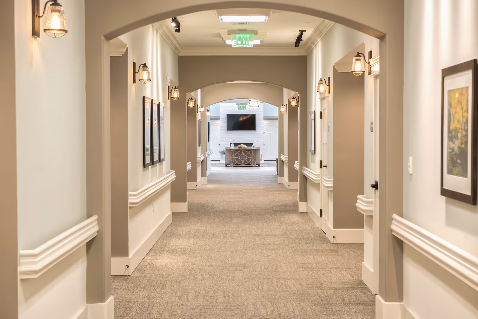 A well-lit hallway in a senior living facility with beige walls, carpeted floor, wall-mounted lights, framed pictures, and an exit sign at the far end above a doorway leading to a room with a TV and a decorative console table.