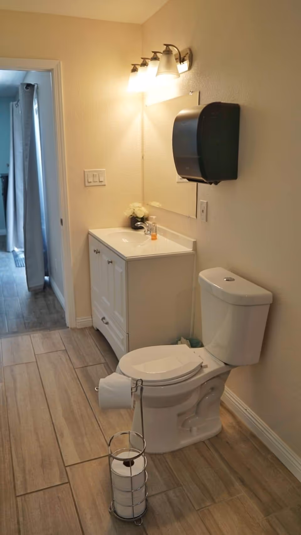 A clean bathroom with a white toilet, a white vanity with a sink, a mirror above the sink, and a wall-mounted black paper towel dispenser. There is a metal stand holding extra toilet paper rolls on the floor. The bathroom has beige walls and wood-look tile flooring. A doorway leads to another room with curtains visible.