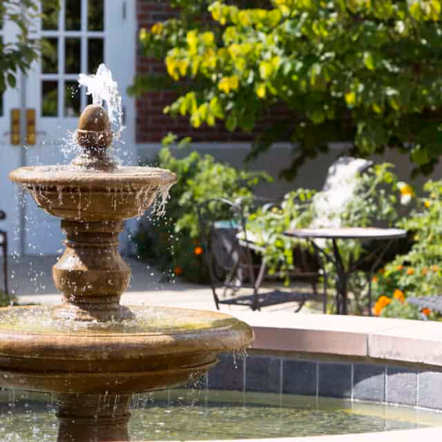 A stone water fountain with water flowing from the top tier into a circular basin, surrounded by greenery and outdoor seating including a table and chairs, with a building entrance visible in the background.