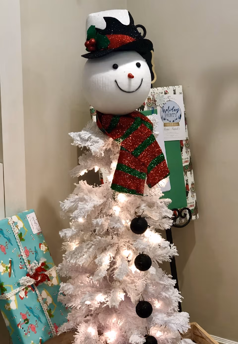A white artificial Christmas tree decorated with white lights and black ornaments, topped with a snowman head wearing a black and white hat with holly and a red and green striped scarf. Wrapped presents are placed beside the tree, and a holiday party invitation is visible in the background.