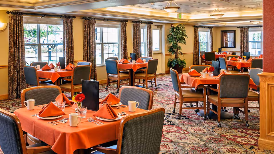A dining room with multiple tables covered in orange tablecloths and neatly arranged with orange napkins, white mugs, glasses, and silverware. The room has large windows with patterned curtains, a patterned carpet, and a potted plant near the back wall.