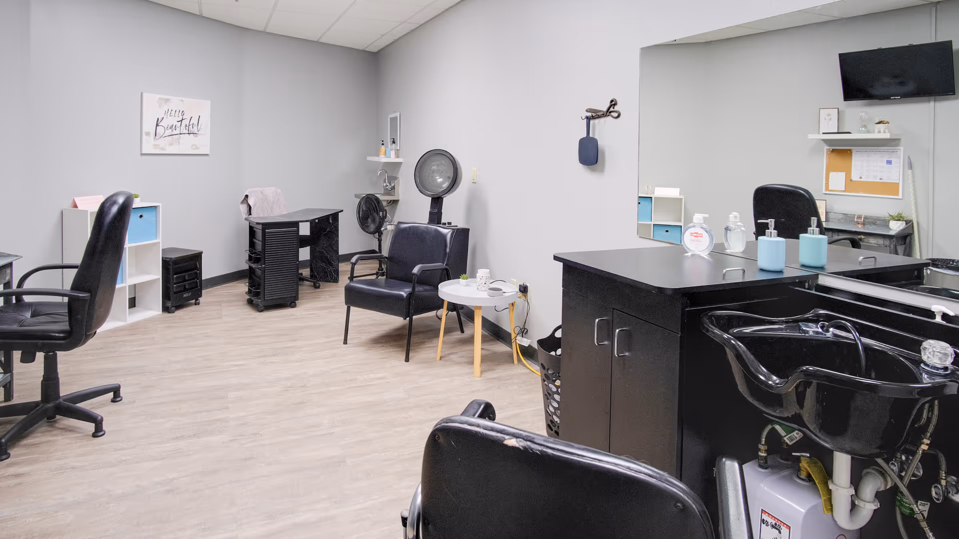 Interior of a senior living facility hair salon with black salon chairs, a hair washing station, a small round table with a cup, a hair dryer, and a wall-mounted TV. The walls are painted light gray and there is a sign that says 'Hello Beautiful'.