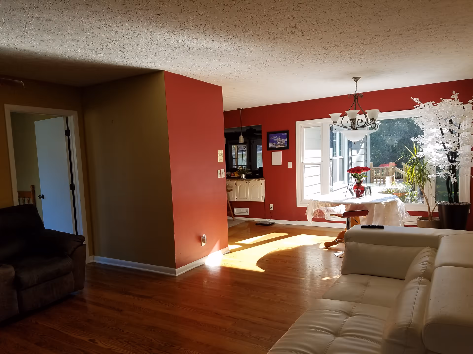 Interior view of a living room and dining area with hardwood floors. The living room has a white leather sectional sofa and a dark recliner chair. The dining area features a round table covered with a white tablecloth, a vase with red flowers, and large windows letting in sunlight. The walls are painted red and beige, and there is a chandelier hanging above the dining table. Potted plants are placed near the windows.