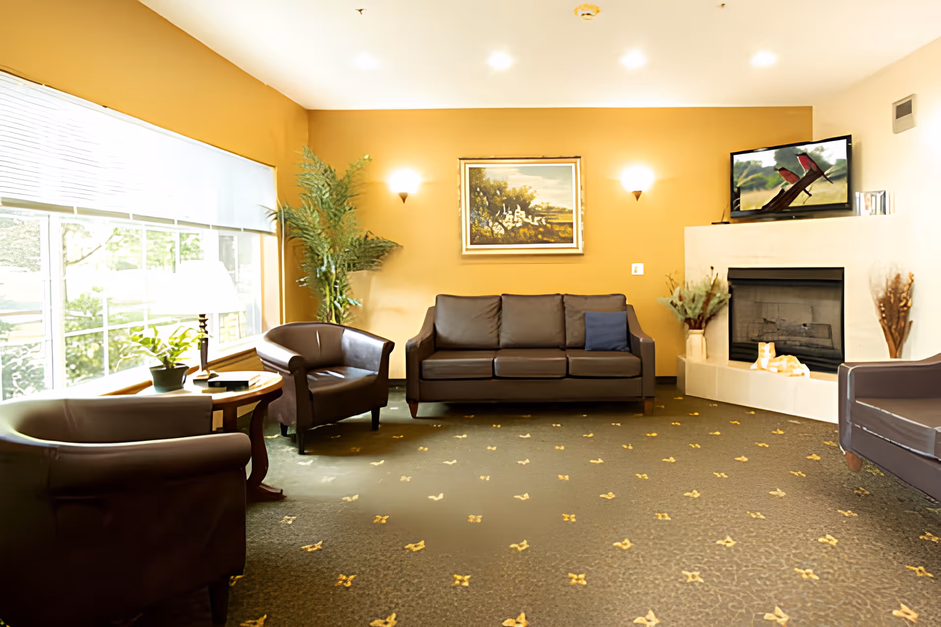 A cozy living room with mustard yellow walls, a large window with blinds, a dark brown leather sofa with a blue cushion, two matching armchairs, a round wooden side table with a lamp and plants, a fireplace with decorative items, and a flat-screen TV mounted above the fireplace.