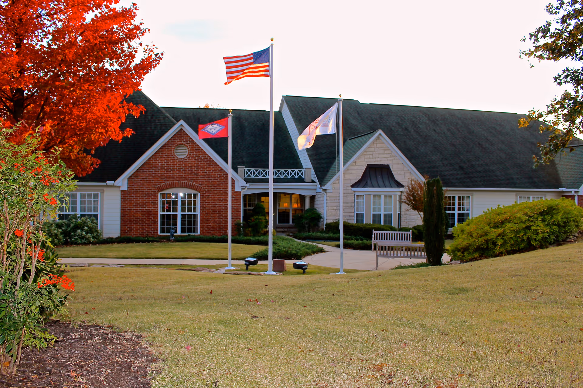 Exterior view of a senior living facility building with a brick and stone facade, three flagpoles with flags including the American flag, a bench near the entrance, and well-maintained landscaping with green grass and trees with autumn foliage.