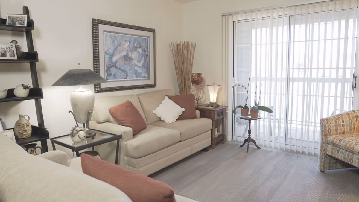 Living room with beige sofas, side tables and lamps, decorative shelving, and a sliding glass door with vertical blinds.