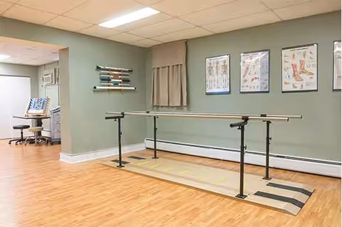 Interior rehabilitation room with parallel bars, open space, wooden floor, and medical posters on walls.
